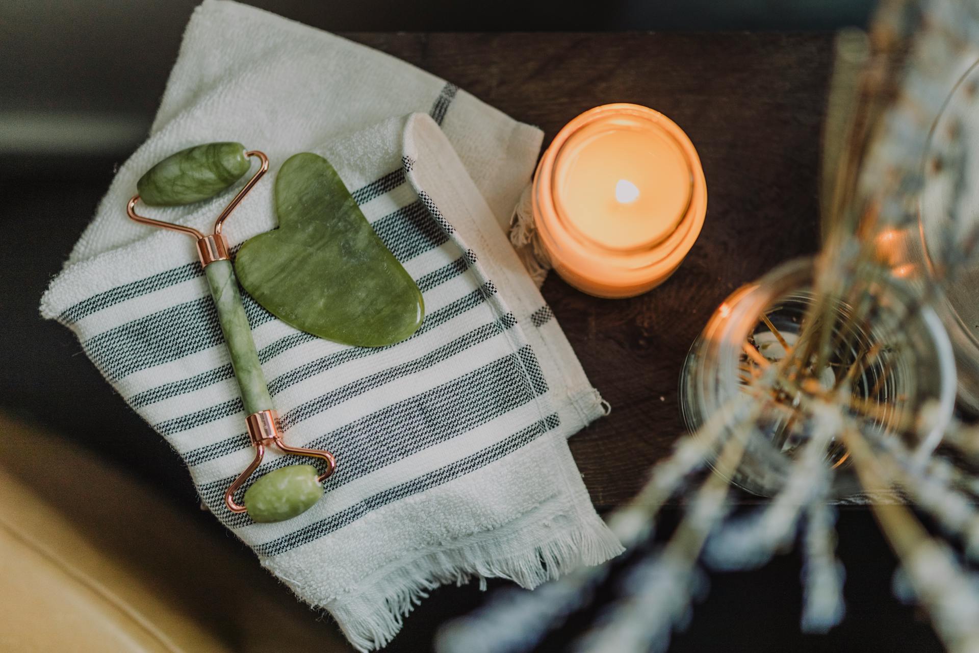 Flat lay of a jade roller, gua sha stone, folded towel, and lit candle on a neutral background